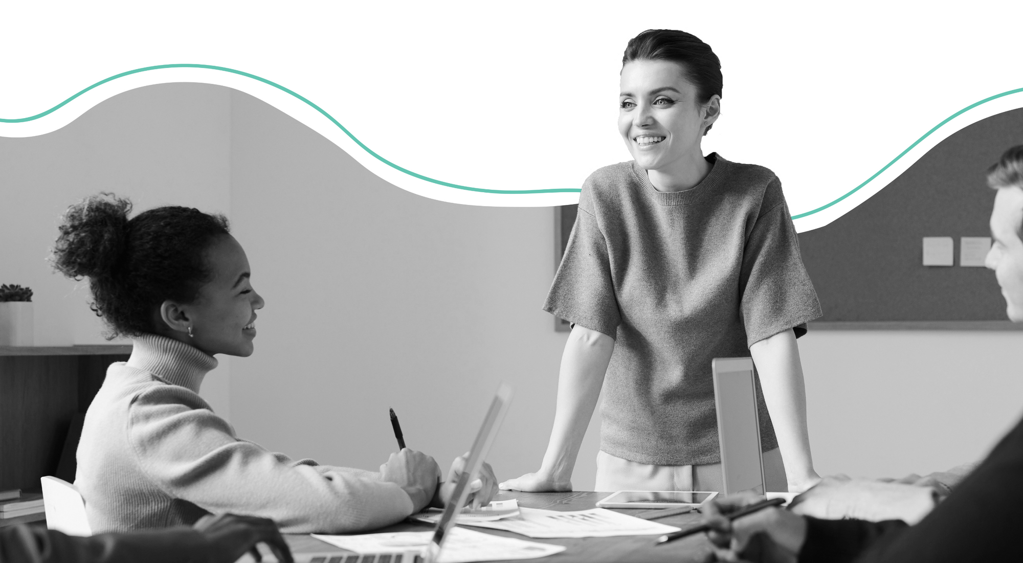 two women at a desk, one standing, one sitting, both smiling and discussing work